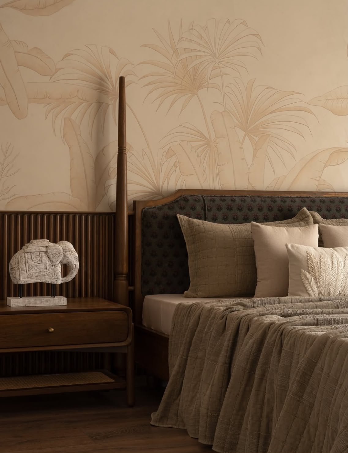 Four-poster bed against a pale palm-leaf mural, with a stone elephant on the bedside table